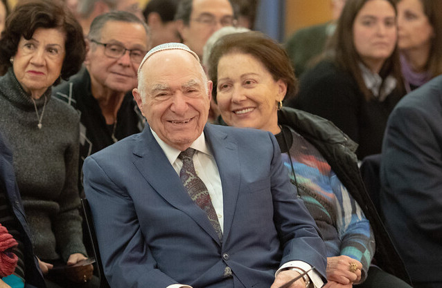 Hazzan Azose smiling in the crowd at Ladnio Day wearing a grey suit, white shirt, and tie. His wife Elisa is next to him.