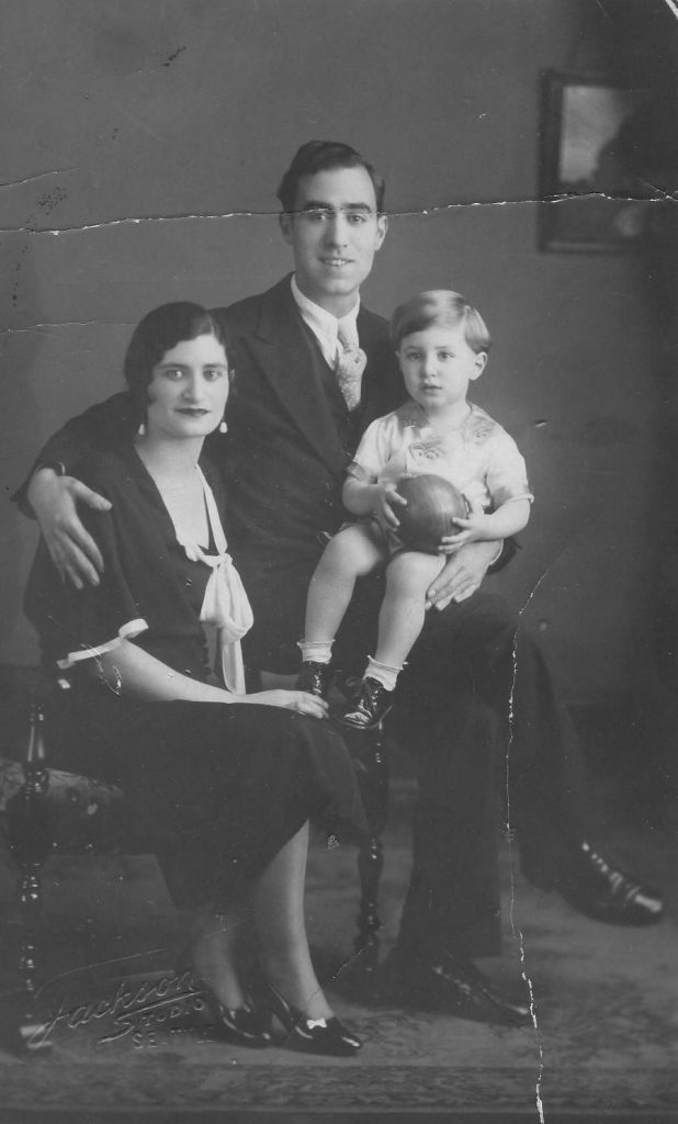 Black and white photo of Hazzan Azose as a child seated on his father's lap, with his mother seated next to them.