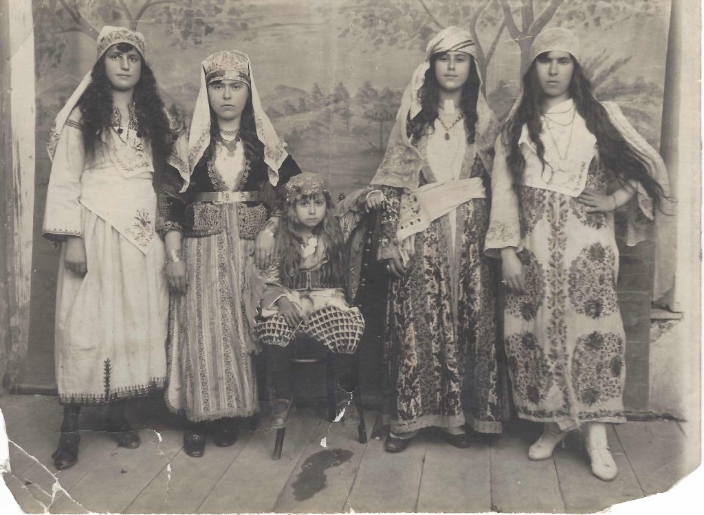 Four women stand dressed in elaborate costumes. They are wearing with dresses and headpieces. A young boy stands at the center.