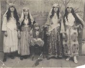 Four women stand dressed in elaborate costumes. They are wearing with dresses and headpieces. A young boy stands at the center.