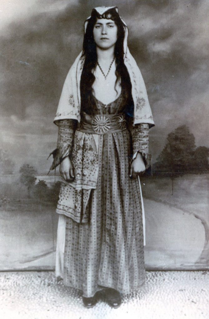 Black and white photo of a woman standing in a purim costume. She wears a long dress with a shawl over top. Her hair is long and black. She wears a headpiece.