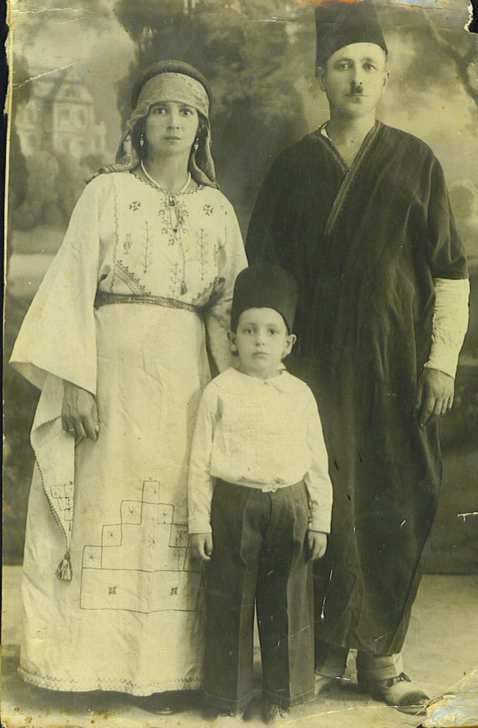 Sepia toned photo of a mother, father, and son in costumes. The mother stands at left and is wearing a long dress with a belt, and a headscarf. The father stands at right and wears a tunic and a fez. Their son stands between them and is wearing slacks, a button down shirt, and a fez.