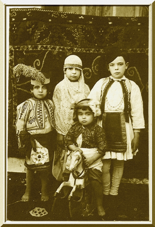 Sepia toned photo of four children in Purim costumes. They are wearing various hats and vests. At center is what appears to be the youngest child sitting on a toy horse.