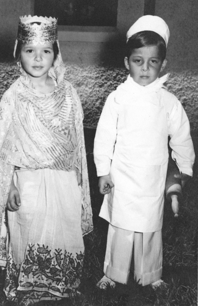 Black and white photo of a young boy and girl in costumes. The girl stands at left and is wearing a queen costume. The boy is at right and is wearing a white tunic, white pants, and a white hat.
