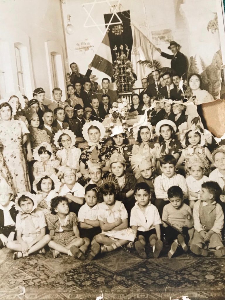 Sepia toned photo of a large group of students seated in Purim costumes. Jewish star hanging in the background. Two men in the background each hold a flag. One is the flag of Mexico.
