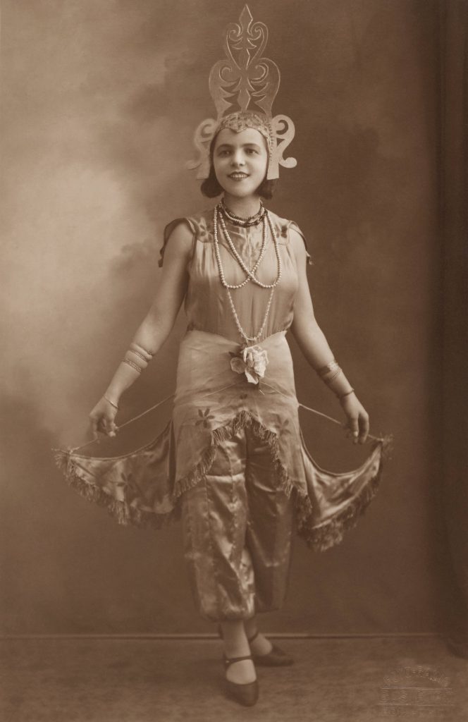 Sepia toned photo of a woman posing in a costume. She is wearing a sleeveless dress and a tall, carved headpiece.