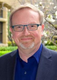 Portrait of Aron Rodrigue. He is wearing a dark grey suit with a royal blue shirt underneath. He has blue glasses and light hair with a goatee. There are cherry blossom trees in the background