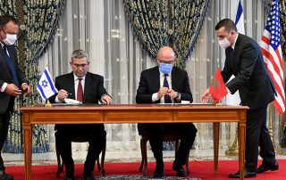 Two officials sit at a wooden table, signing the Israel-Morocco Normalization Agreement, with an Israeli flag on one side and a Moroccan flag on the other