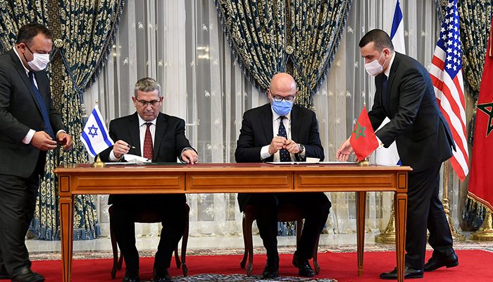Two officials sit at a wooden table, signing the Israel-Morocco Normalization Agreement, with an Israeli flag on one side and a Moroccan flag on the other