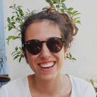 Portrait of Joana Burger. She is wearing sunglasses, a white tshirt, and has her hair in a bun. There are leaves in the background
