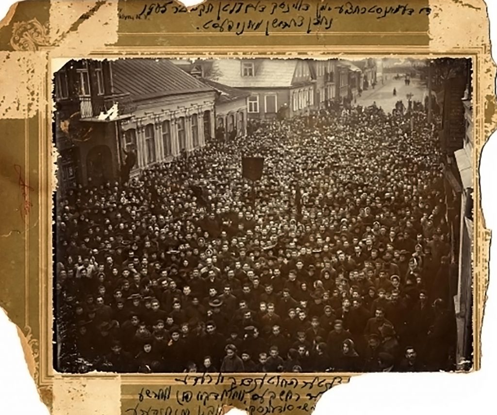 Black-and-white photograph showing hundreds of people gathered in a large town plaza, with handwritten notes in Hebrew around a paper frame