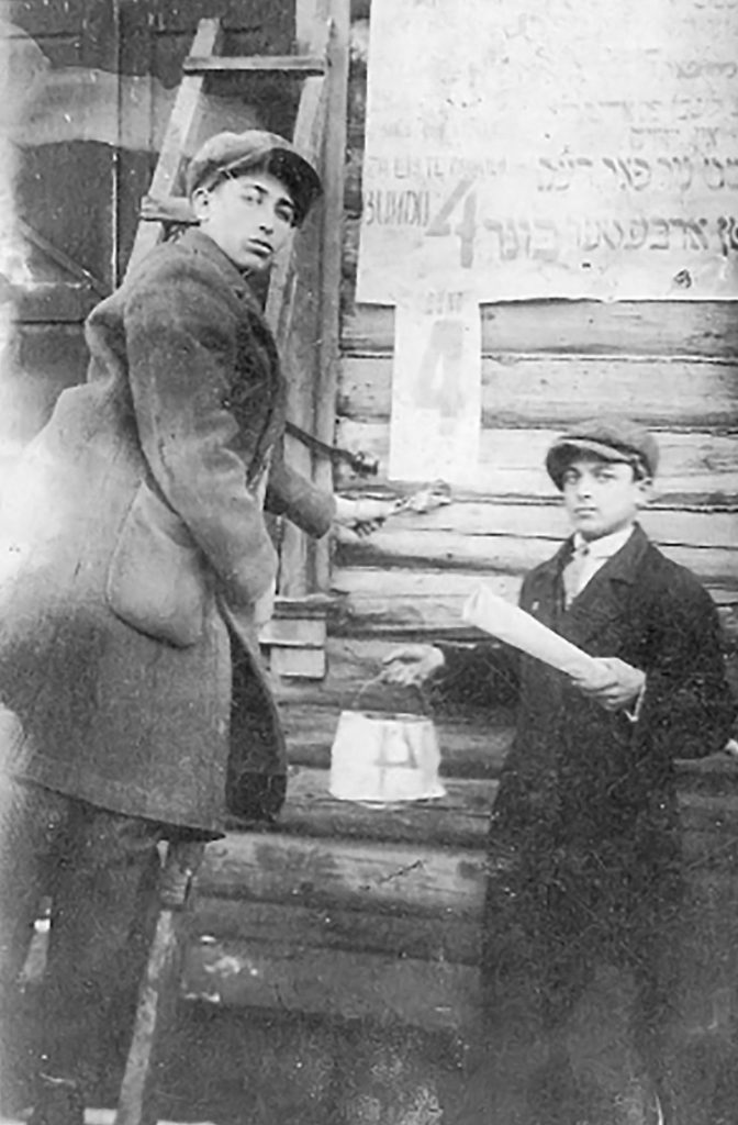 Black and white photo of two boys wearing heavy jackets and hats hang up posters in Hebrew