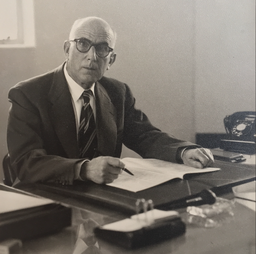 Haim Galante at his desk. He is wearing a suit, tie, and glasses. He is writing in a notebook. 