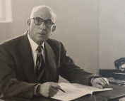 Haim Galante at his desk. He is wearing a suit, tie, and glasses. He is writing in a notebook.