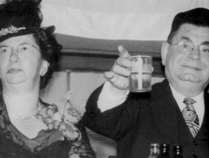Black and white photo of Morris and Esther Scharhon. Morris is at right, wearing a suit and holding up a glass, as if toasting. Esther is on the left and is wearing a hat and a dress, with a boutonniere on her lapel.