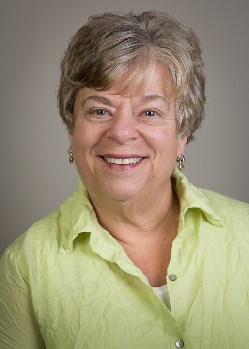 Portrait of Doreen Alhadeff. She is wearing a green button down shirt and smiling. She has short, grey hair. The background is grey.