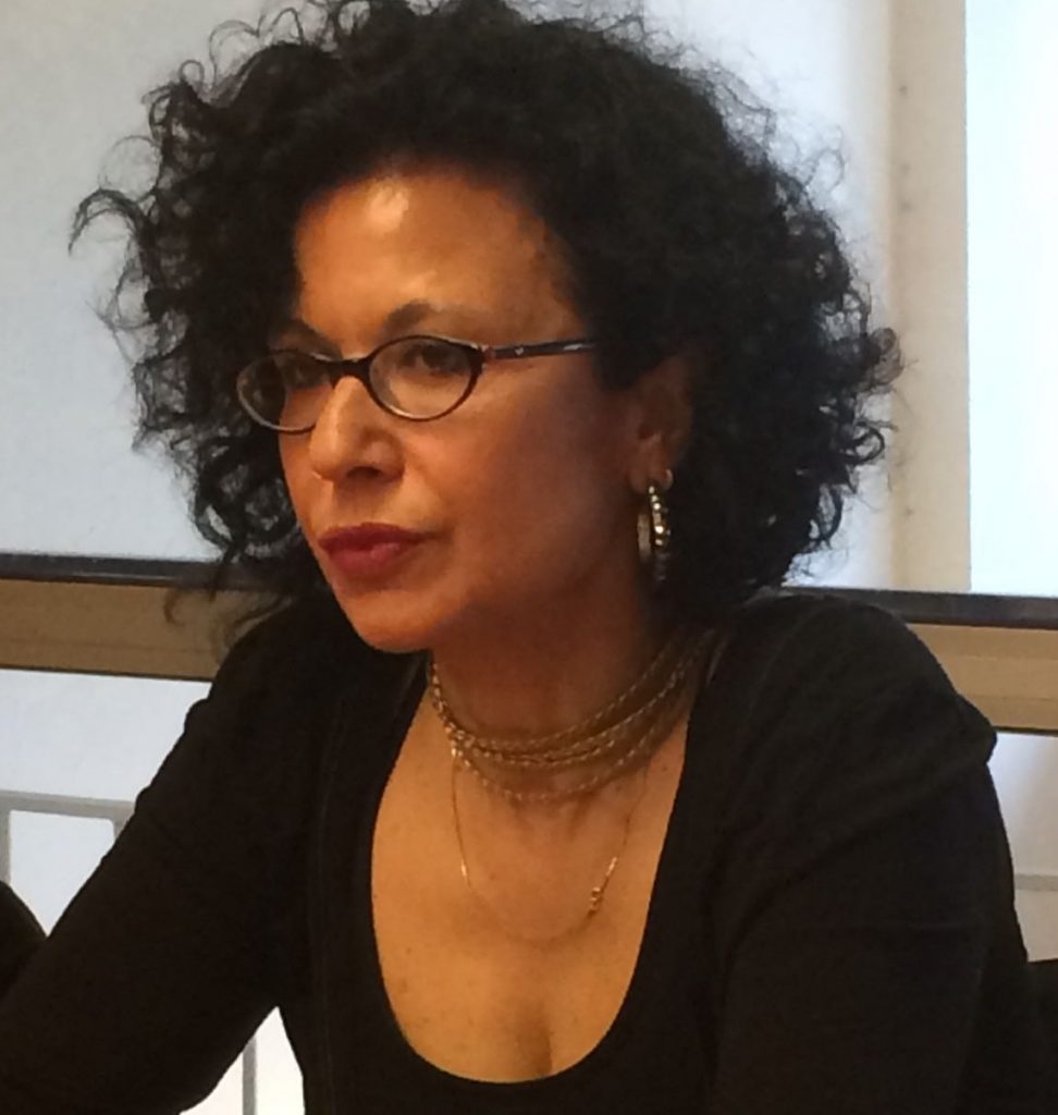 Headshot of Rasel Maseri. She is wearing a black shirt and glasses and has dark curly hair. There are windows in the background.