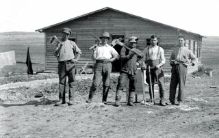 Surrounded by desert and sun, 5 Jewish settlers pose for a photo in front of a new wooden building with their handheld construction tools