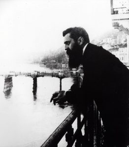 B&W photo of Herzl, during the First Zionist Congress, watches on the metal railing, turns his side to the camera and looks away. In the background is a bridge over the river and the houses of the city.