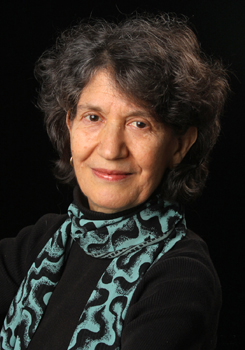 portrait of Jane Mushabac smiling on a black background, wearing a black shirt and a teal and black scarf with a funky pattern