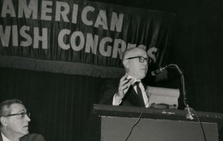 Black & white photo of Leo Pfeffer speaking at the National Convention of the American Jewish Congress in 1966. Seated is Shad Polier.