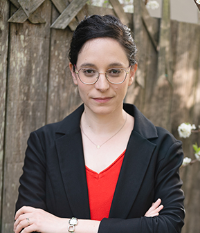 Portrait of Masua Sagiv smirking with arms crossed, wearing red blouse, black blazer, and large circular glasses with a wooden fence in background