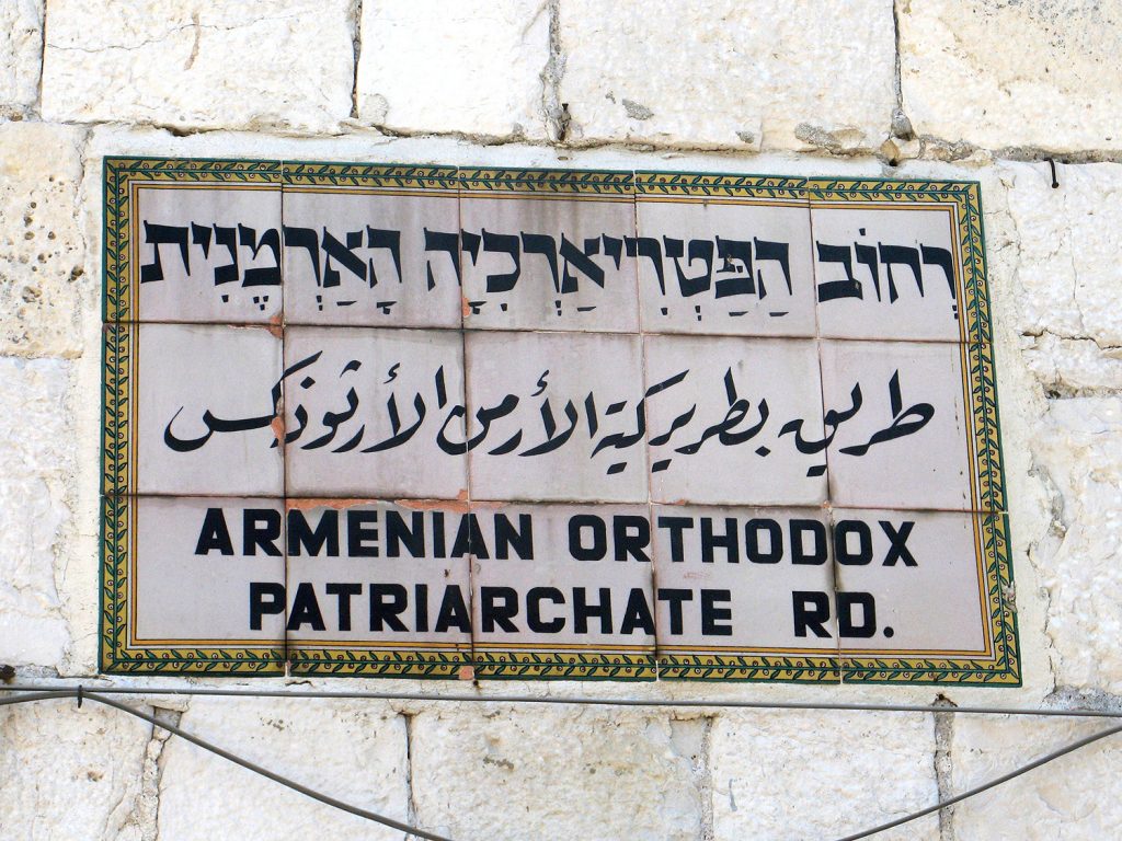 Tile sign embedded in a stone wall, with writing in Hebrew, Arabic, and English: "Armenian Orthodoc Patriarchate Rd."