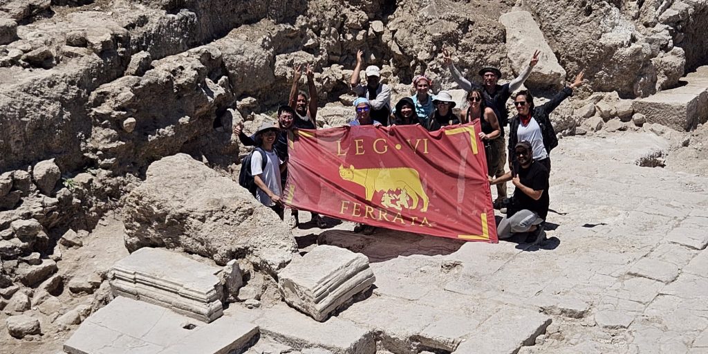 Students and staff hold up a Legio 6 flag at their excavation site