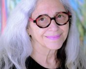 portrait of artist Linda Dayan Frimer smiling with bright white hair, pink lips, and artsy glasses