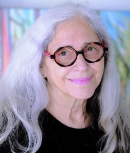 portrait of artist Linda Dayan Frimer smiling with bright white hair, pink lips, and artsy glasses
