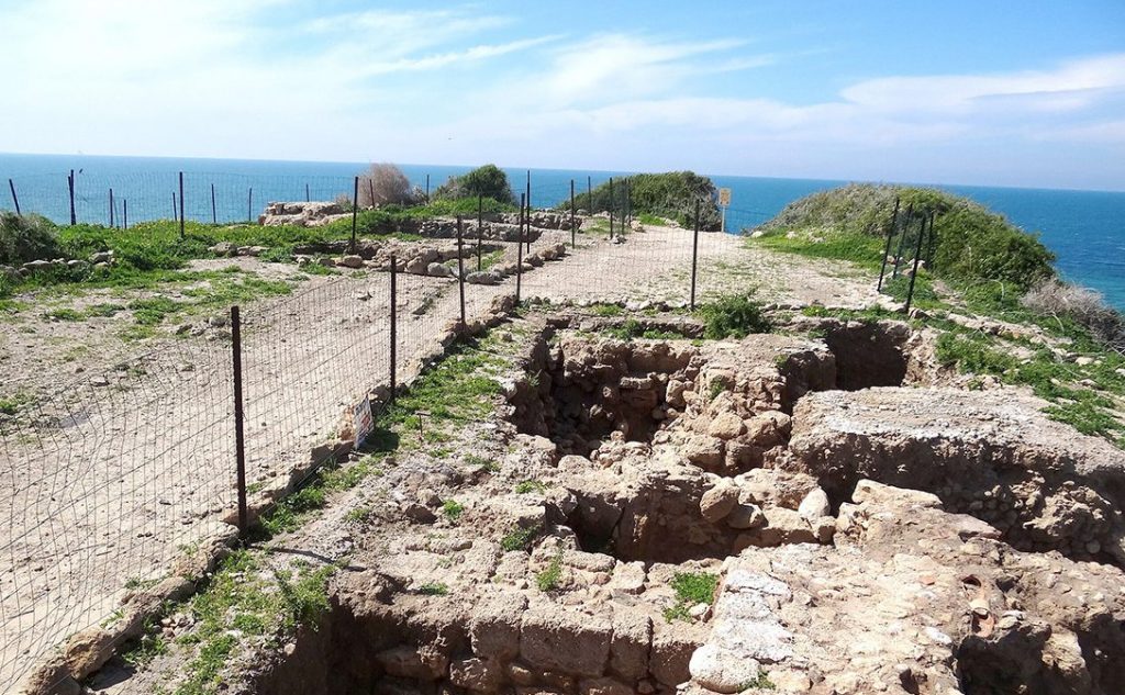 Ancient stone structures below ground in a fenced-off area, sunny sky and sea visible