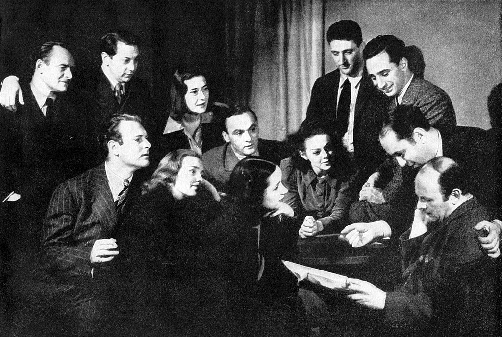Black-and-white photo of formally dressed men and women (in suits or buttoned-up jackets) grouped around a man reading aloud from a script