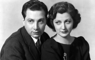 Black-and-white photo showing a glamorous man and woman sitting close together, both wearing dark formal clothing