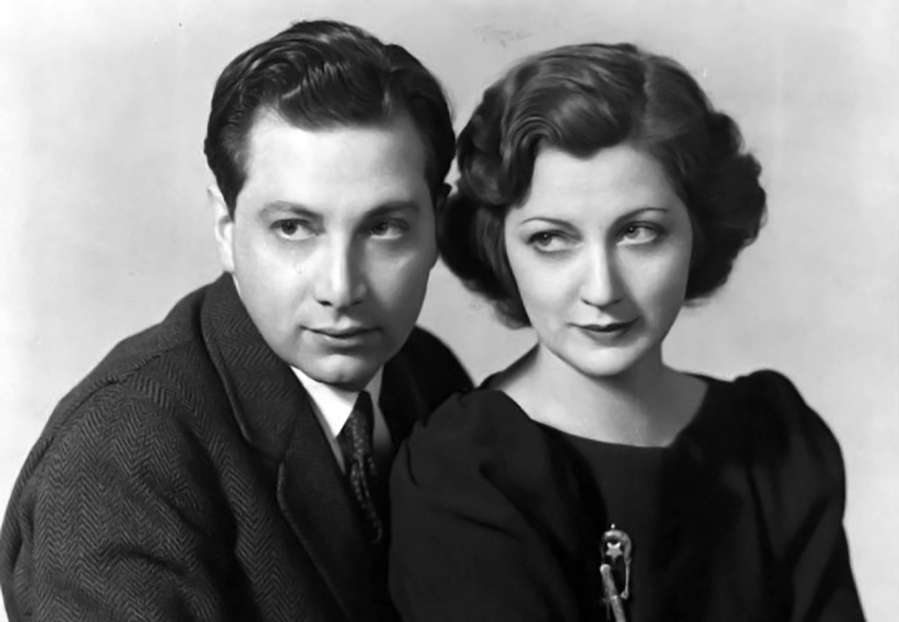 Black-and-white photo showing a glamorous man and woman sitting close together, both wearing dark formal clothing