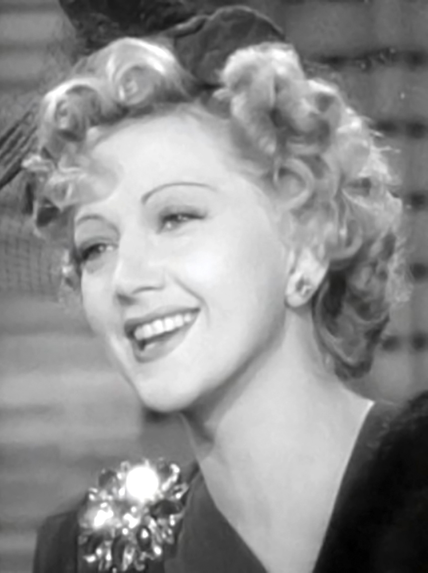 Black-and-white screenshot of a youthful, smiling Stella Adler in glamorous makeup, wearing a small, stylish hat atop curled blond hair
