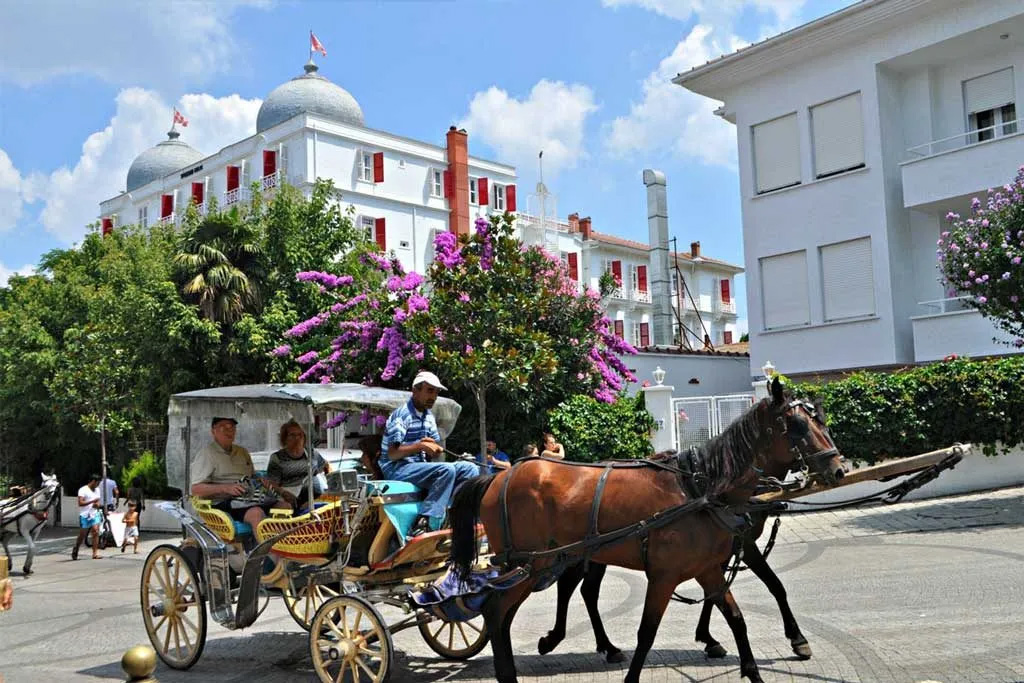 People ride in an old-fashioned horse-drawn carriage along wide cobblestone street, with historic buildings in the background