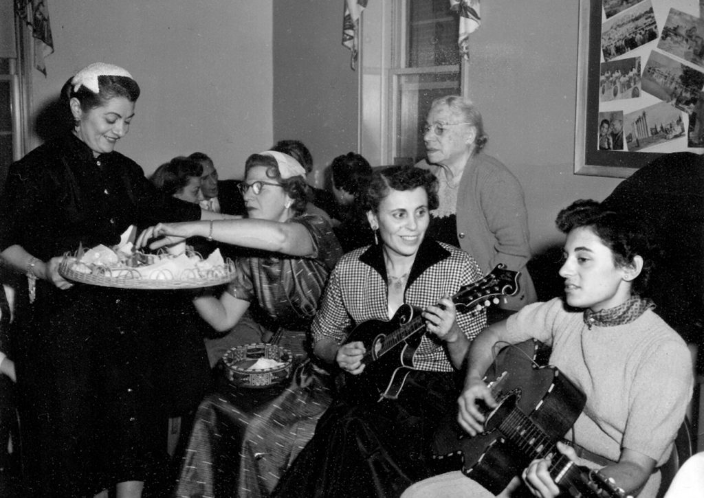 Black-and-white photo of a party with women seated on couch, two laying the guitar