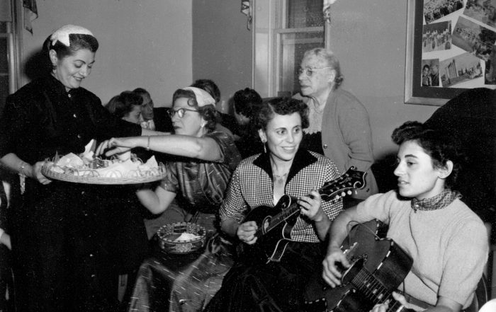 Black-and-white photo of a party with women seated on couch, two laying the guitar
