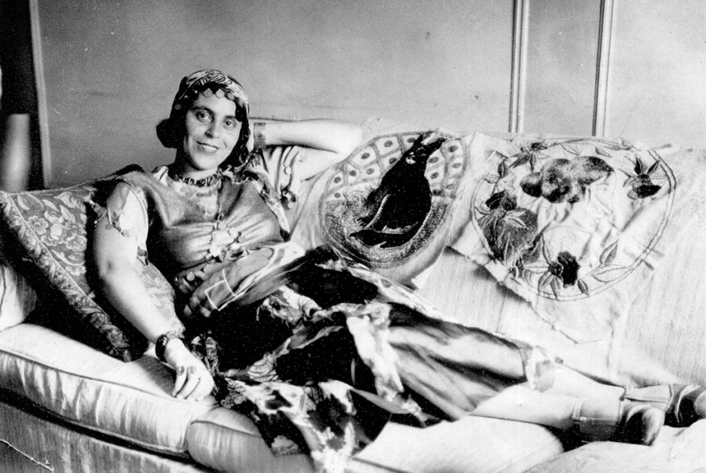 Black-and-white photo of woman in Mediterranean outfit reclining on couch and smiling