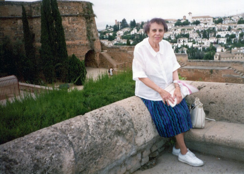 Older woman in white shirt and skirt seated on stone rampart, medieval castle and Spanish city visible in background