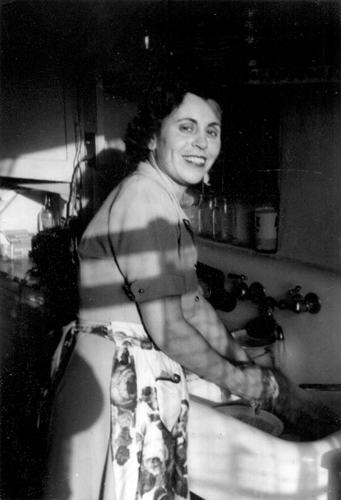 Black-and-white photo of woman in dress and apron smiling at sink