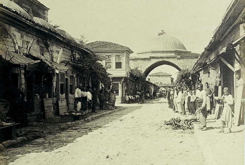 Black-and-white photo showing a cobblestone road leading to an arched bridge with small dome. Boys and men in fezes, white shirts, and baggy trousers look at the camera.