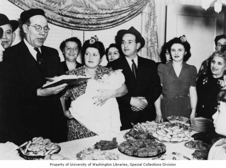 Black-and-white photo showing man in suit singing with open book; mother holds baby at side, friends and family around, with table covered with Sephardic treats in front