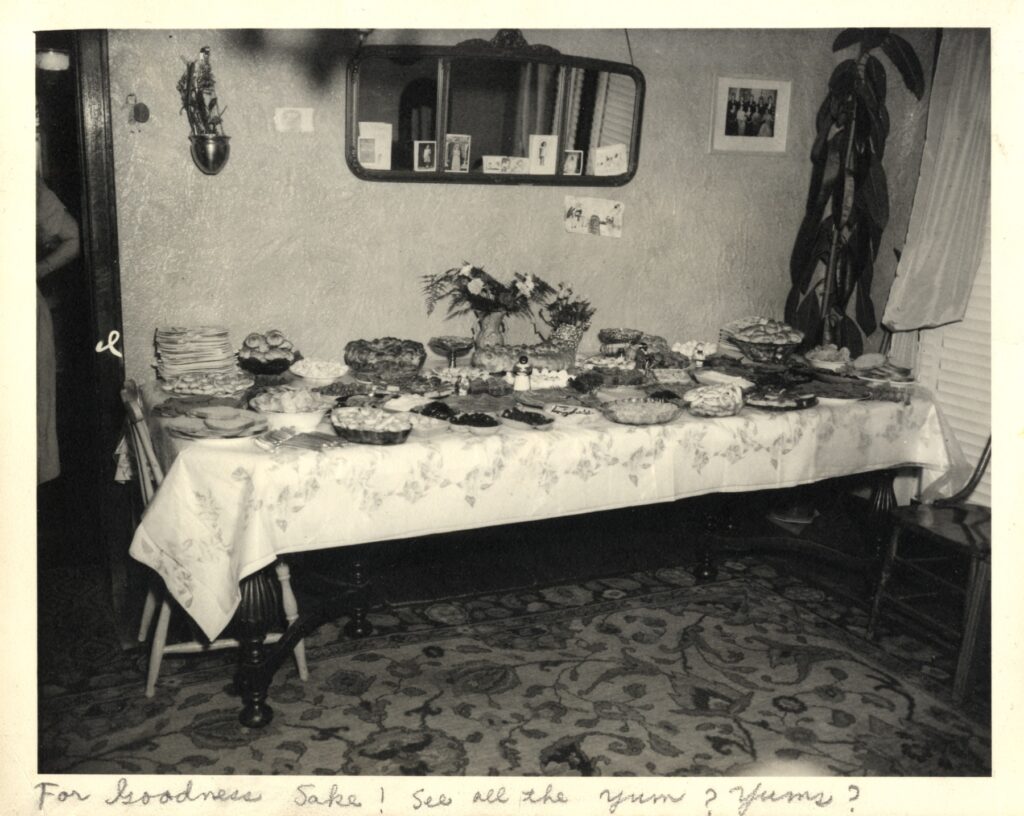 Black and white photo showing a long table with ornate tablecloth fully covered with dishes of treats, a long challah and bouquets of flowers in the center
