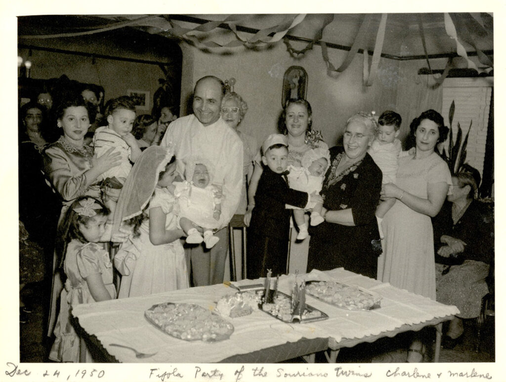 Black-and-white photo of happy family members dressed in formalwear, including five young children, holding up crying babies in white dresses and fluffy caps