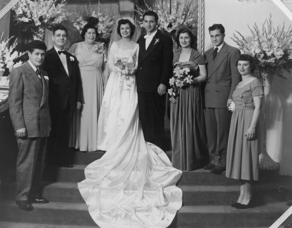 Black and white photo of family members in formalwear grouped around bride and groom on indoor stairs, floral arrangements all around