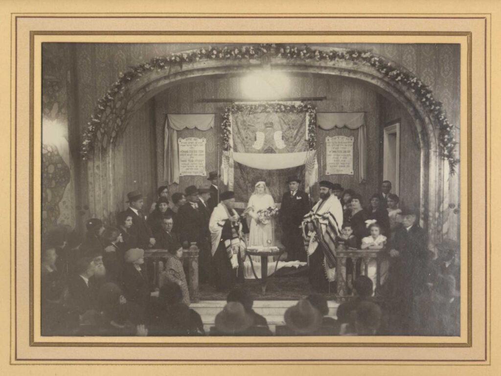 Black and white photo of wedding ceremony in crowded large space with bride and groom and two rabbis wearing hats and prayer shawls in the middle
