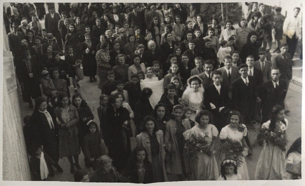High-angle black and white photo of a group of 75 or more people of all ages in formalwear crowding around the bride and groom on a city street