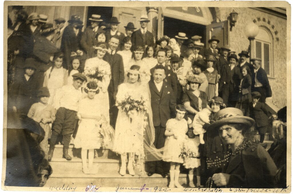 Black-and-white photo of large group surrounding bride and groom outdoors on synagogue steps