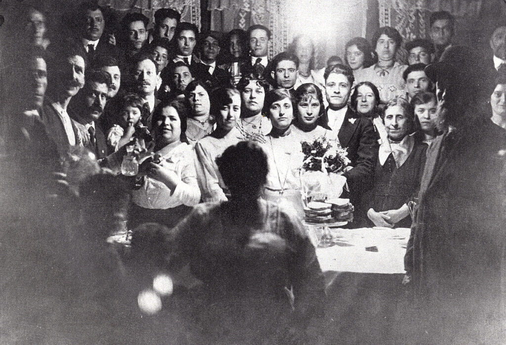 Black-and-white photo of large group of people crowded around bride and groom at home, with table and bouquet in the middle